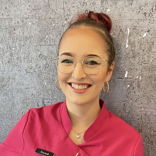 Sarah im pinken Kosmetikkittel lächelt vor grauer Wand